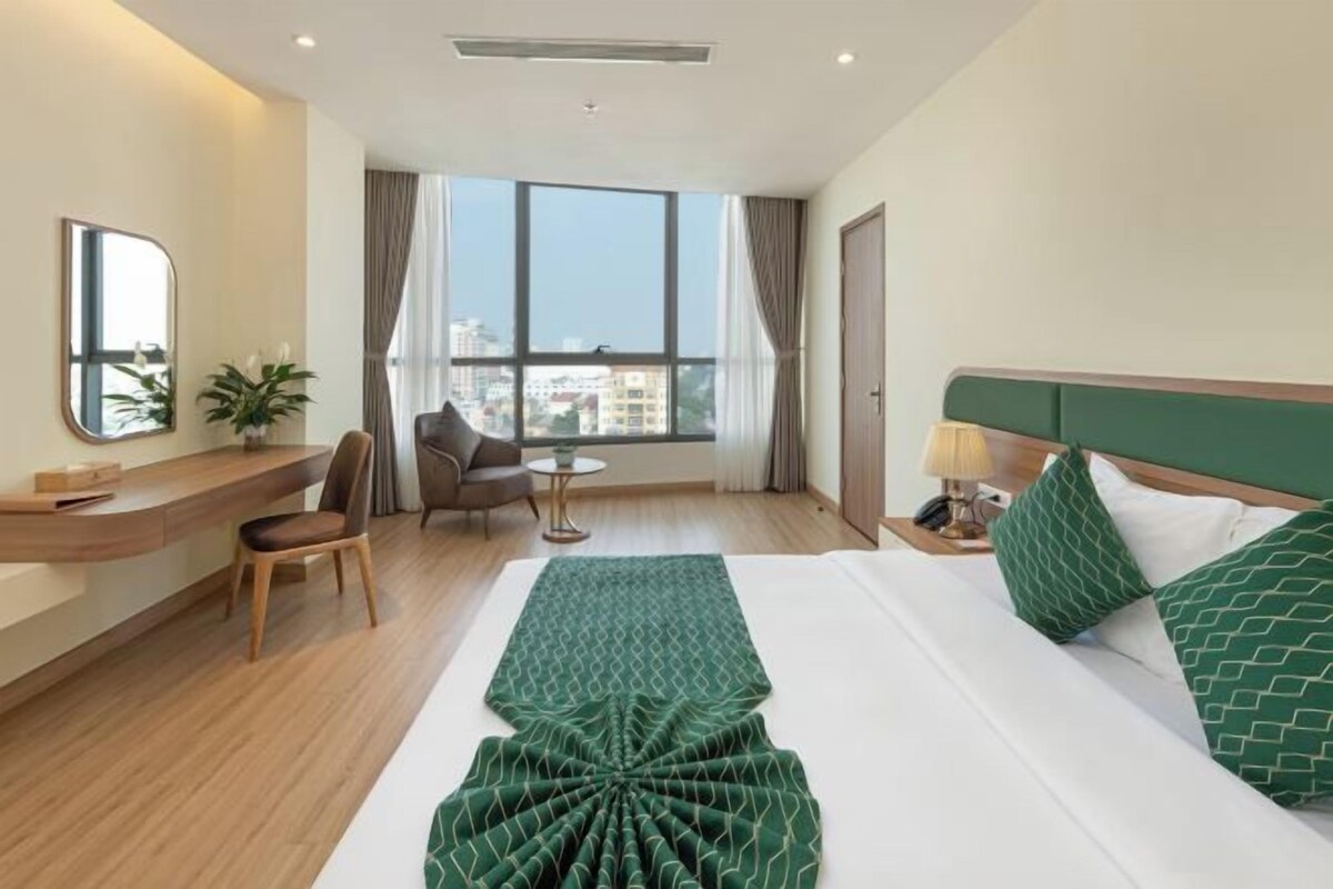 A hotel room with a large bed, a desk, a chair, a small table, and a view of the city through large windows.