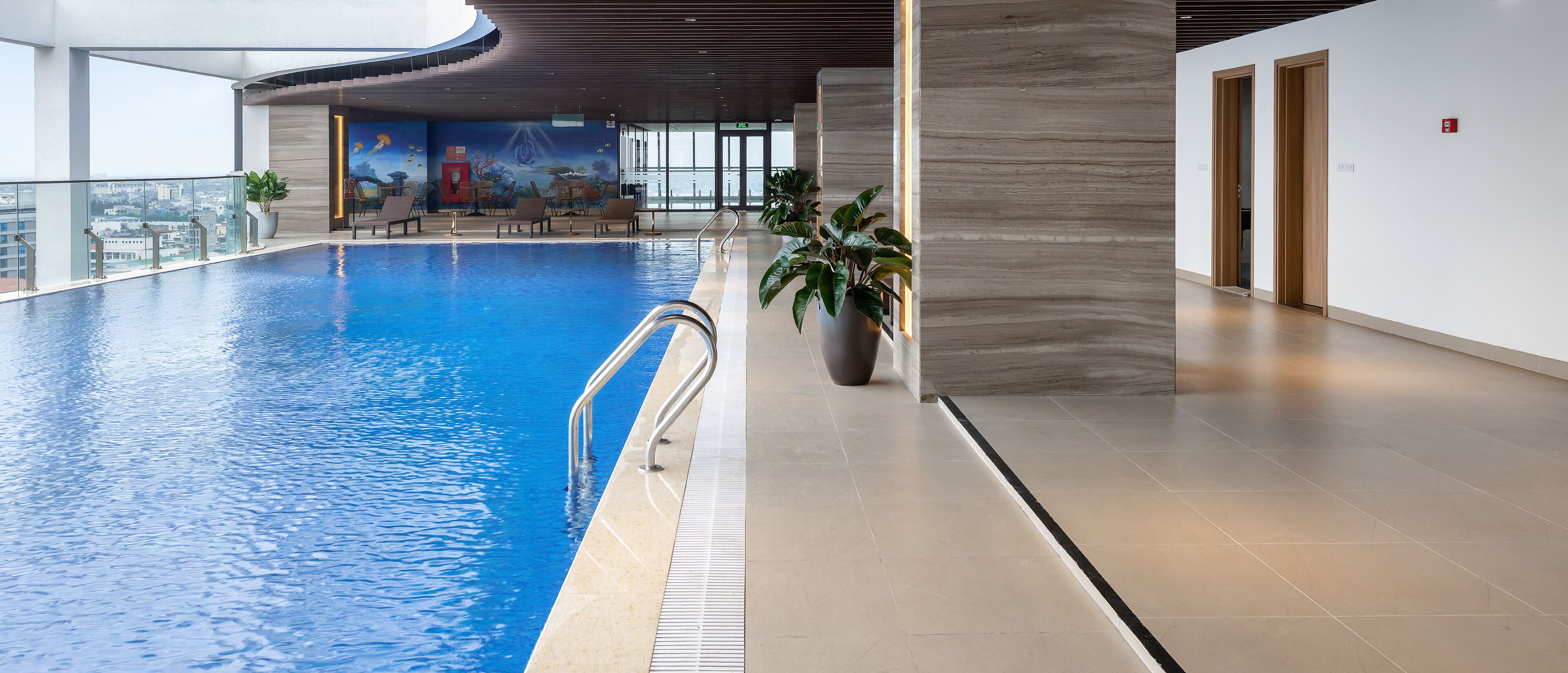 A modern indoor swimming pool with clear blue water, lounge chairs, and a mural on the wall.