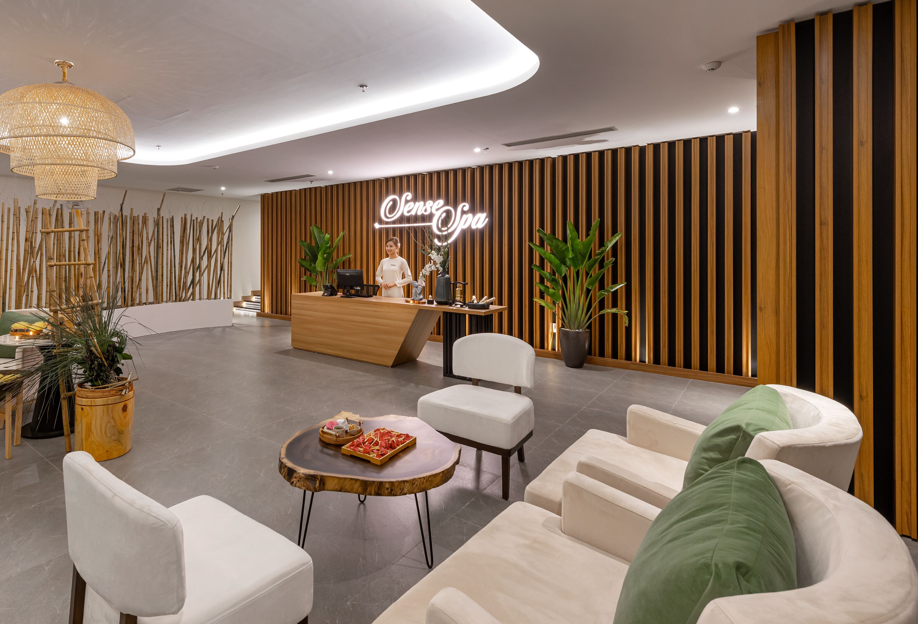 A modern reception area with a wooden reception desk, white chairs, a round table with a tray of snacks, and a large hanging light fixture.