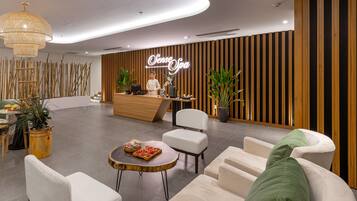 A modern reception area with a wooden reception desk, white chairs, a round table with a tray of snacks, and a large hanging light fixture.