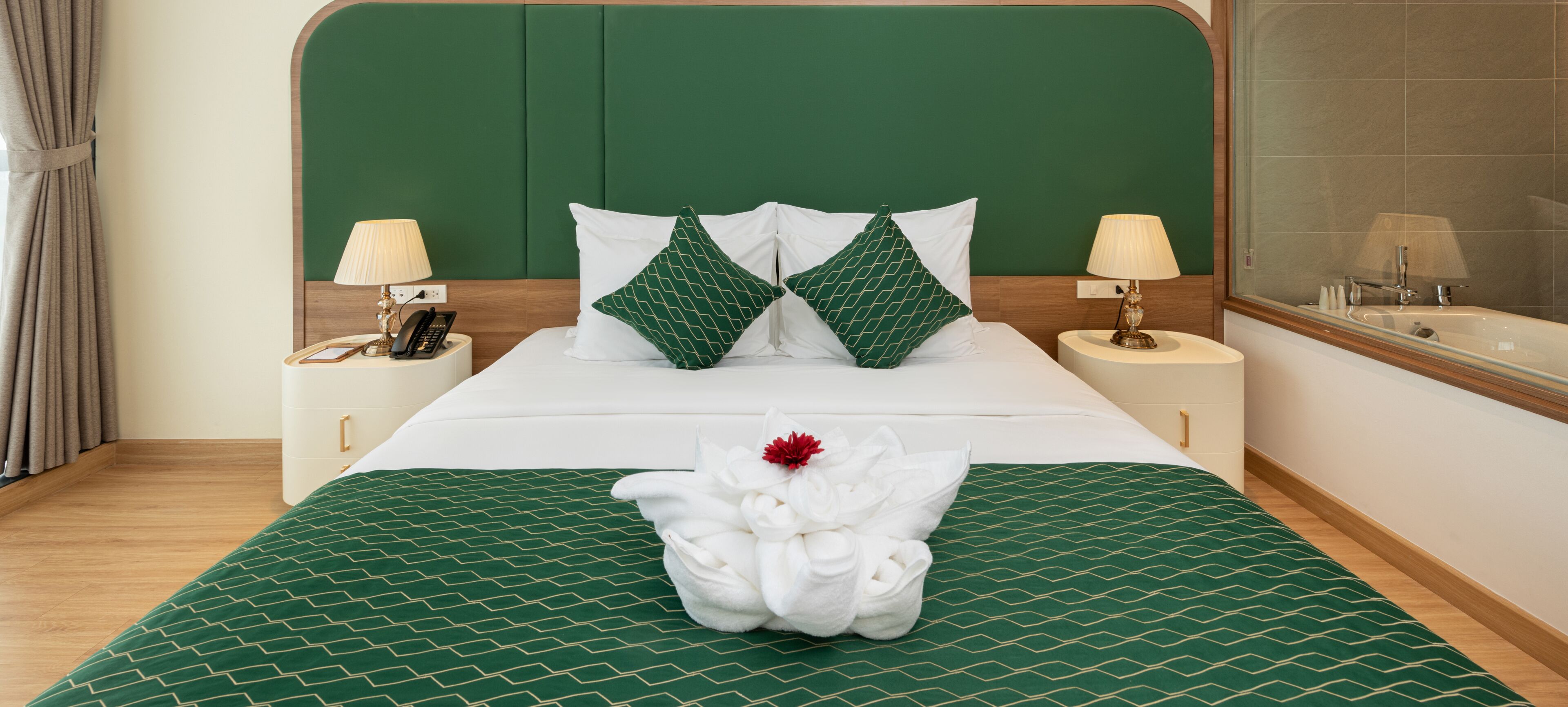 Una cama bien tendida con ropa de cama verde estampada, dos lámparas de noche y un baño visible a la derecha.