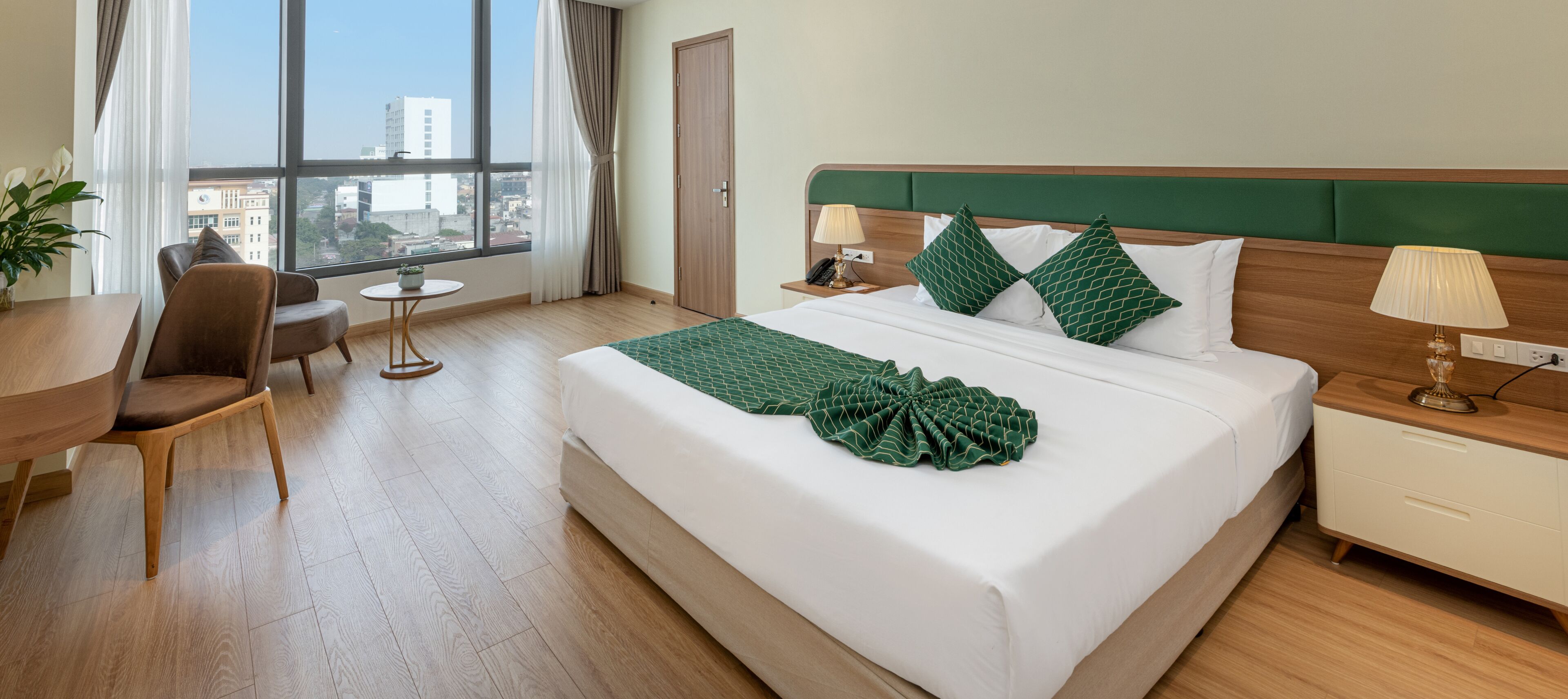 A hotel room with a large bed, a desk, a chair, a small table, and a view of the city through large windows.