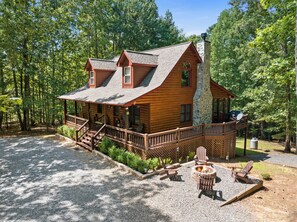 A log cabin with a stone chimney, a deck with Adirondack chairs, and a fire pit.