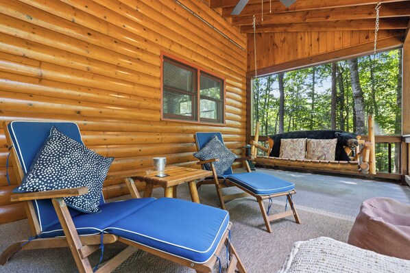 A log cabin with two blue lounge chairs, a wooden table, and a view of the forest.