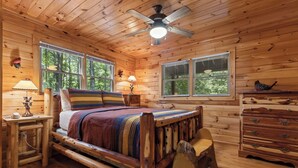 Une chambre dans une cabane en bois, avec un lit, deux fenêtres, un ventilateur de plafond et une commode.