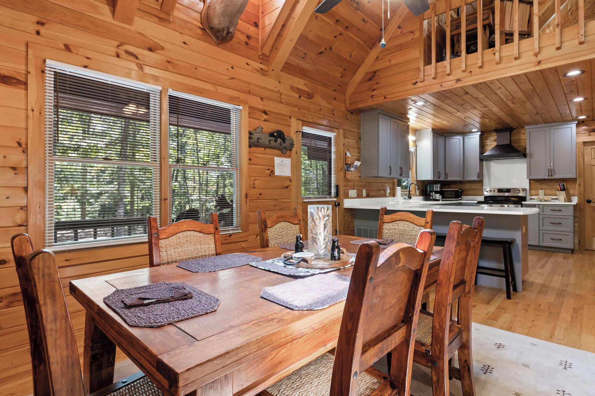 A wooden cabin interior with a dining table, chairs, kitchen area, and large windows.