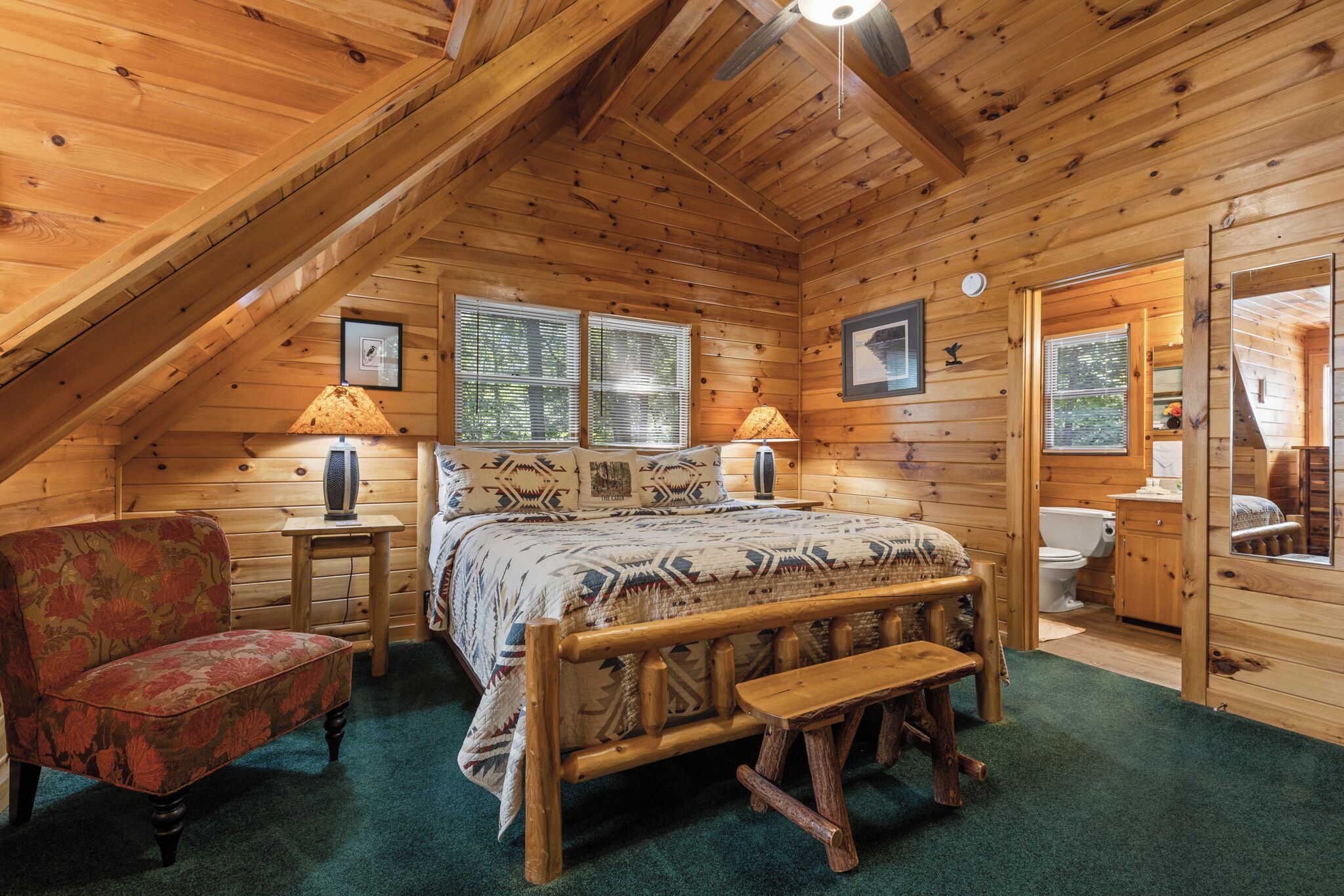 Une chambre mansardée chaleureuse avec un plafond en pente en bois, un lit avec une literie à motifs, une chaise, une petite table et une fenêtre avec des stores.