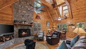 L’intérieur chaleureux d’un chalet avec une cheminée en pierre, des murs en bois, un ventilateur de plafond et un téléviseur.