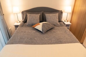Cama bem arrumada com manta cinza e duas almofadas, com luminárias de cabeceira em mesas de cabeceira brancas.