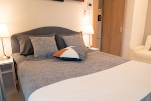 Cama bem arrumada com roupa de cama cinzenta e uma combinação de almofadas cinzas e brancas. Há duas mesas de cabeceira com luminárias, uma porta de madeira e uma cadeira branca no quarto.