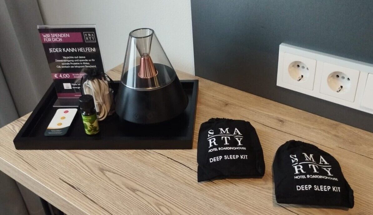 A hotel room with a wooden nightstand, a black tray with a glass candle, a bottle of lotion, a small card, and a black hat with "SMARTY HOTEL BOARDINGHOUSE DEEP SLEEP KIT" text.