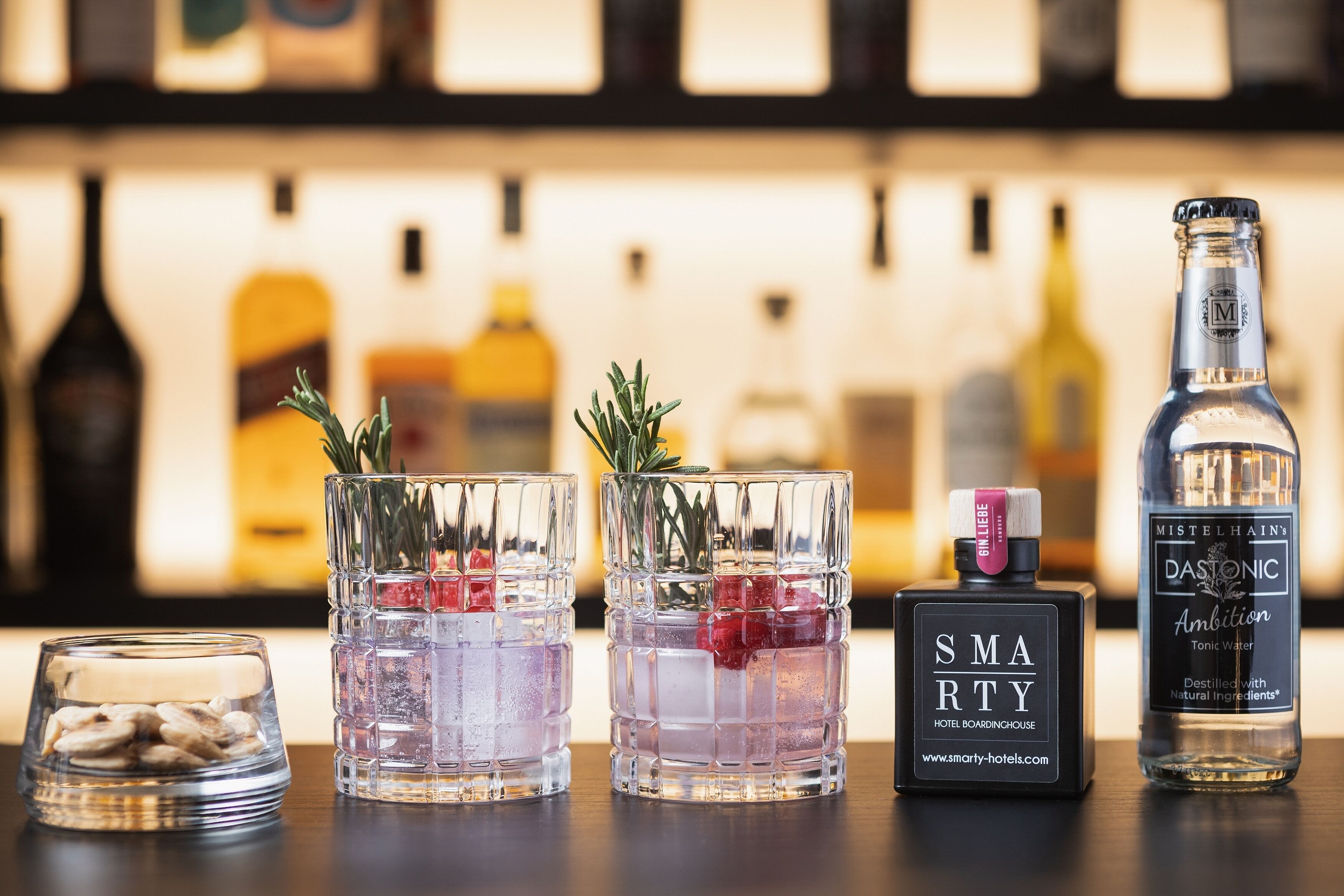 Tres vasos con cubitos de hielo y ramitas de romero, una botella de tónica Mistletoe Ambition y una botella de colonia Smartery Hotel Boardinghouse sobre una barra.