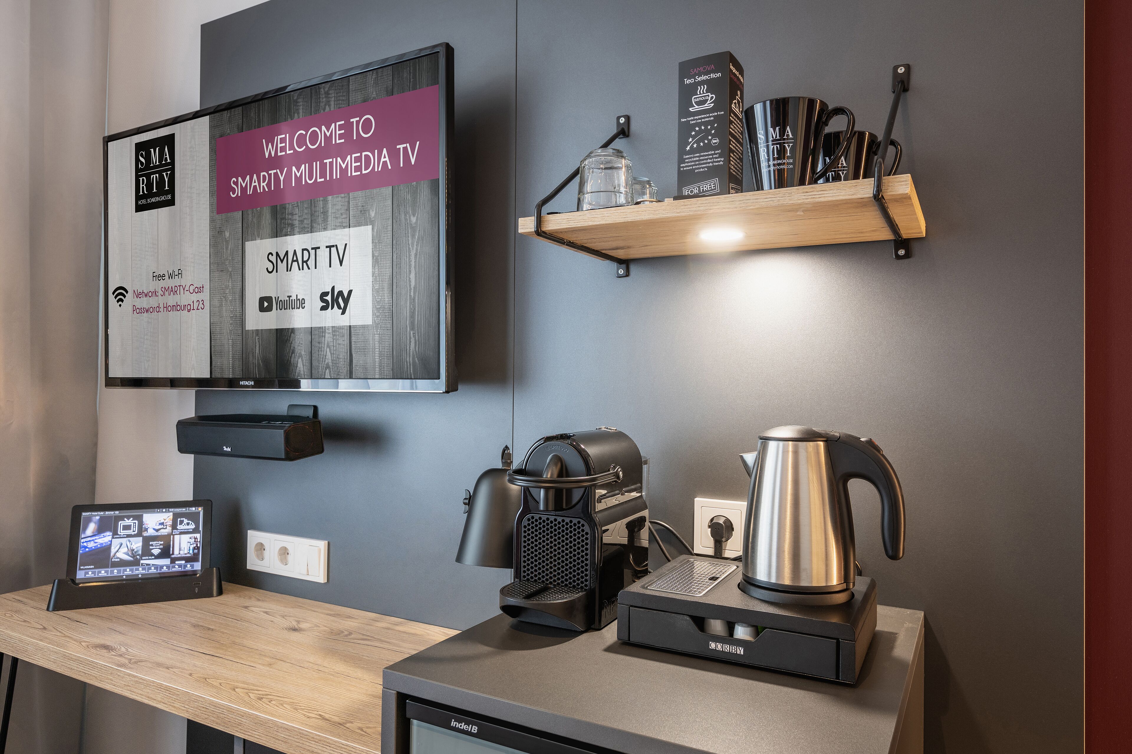A modern hotel room with a flat-screen TV displaying a welcome message, a coffee machine, a kettle, and a small table with a tablet.