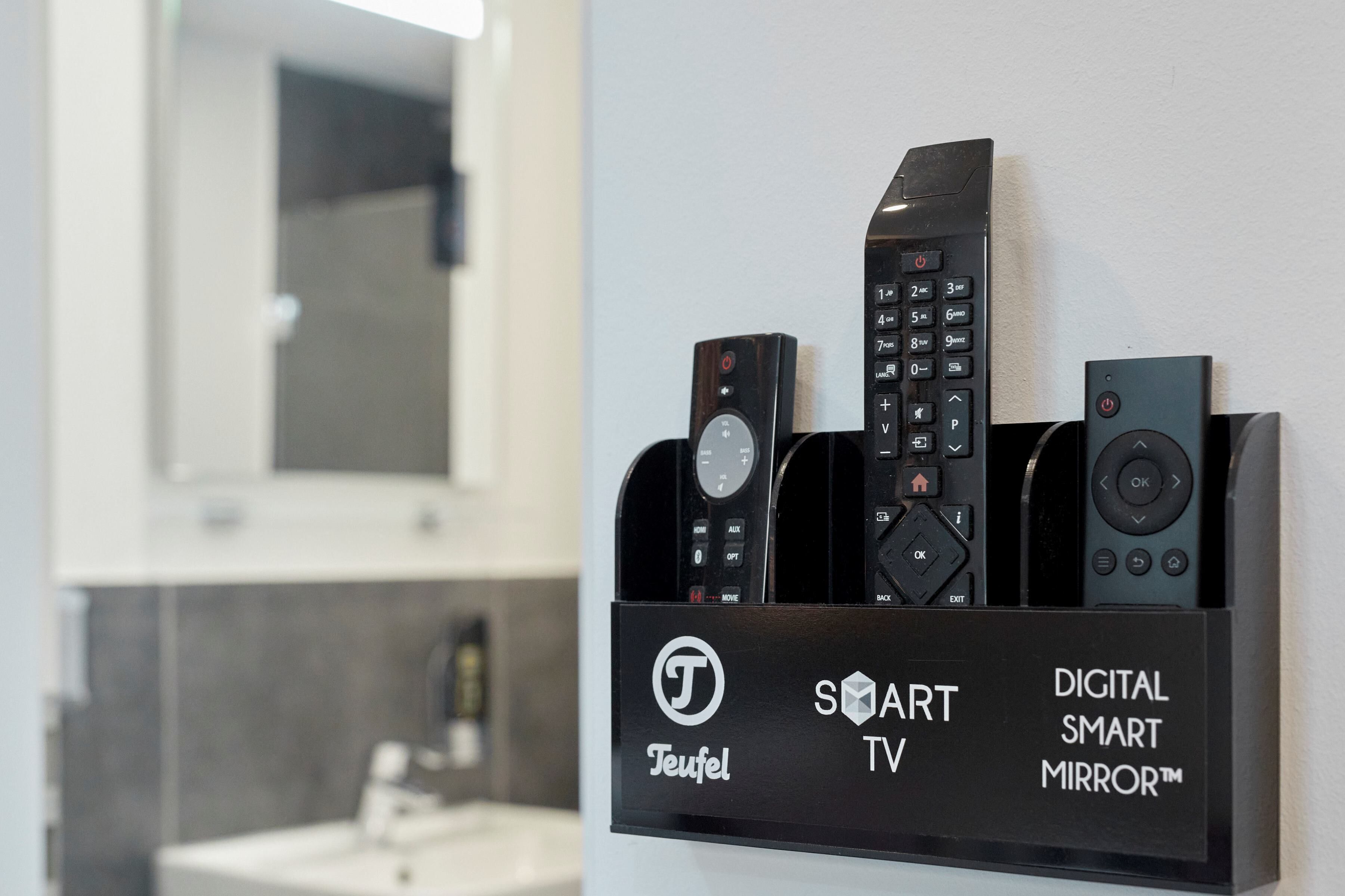 Un mueble de pared para televisor con varios controles remotos, un espejo y un letrero que indica que es un televisor Teufel Smart con un Espejo Inteligente Digital.