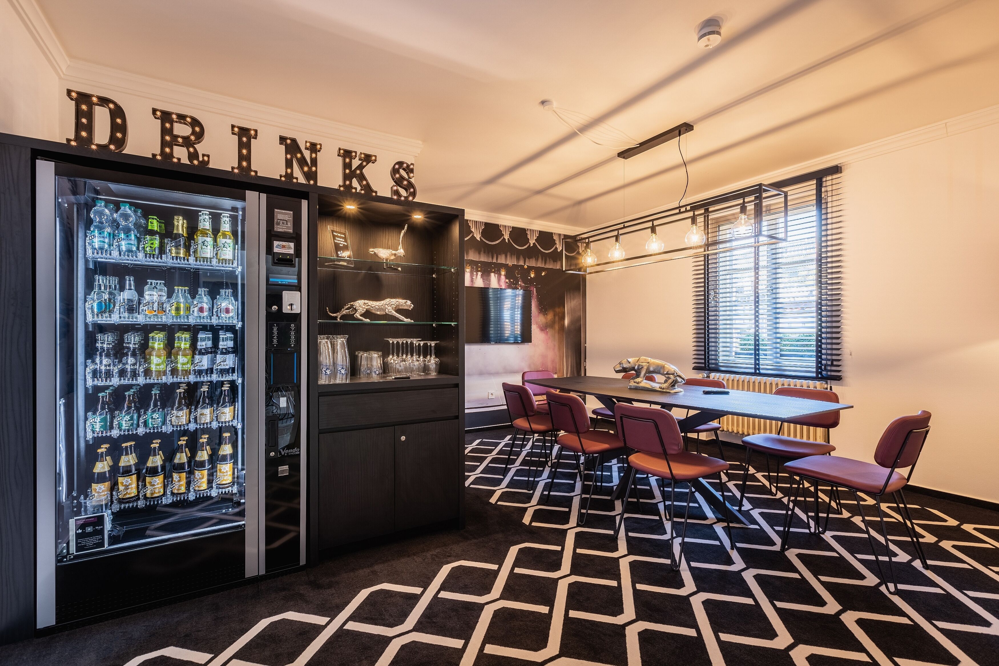 A modern bar area with a drinks vending machine, a dark wooden bar counter, and a seating area with a patterned carpet and a table with chairs.