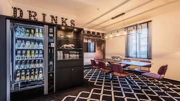 A modern bar area with a drinks vending machine, a dark wooden bar counter, and a seating area with a patterned carpet and a table with chairs.