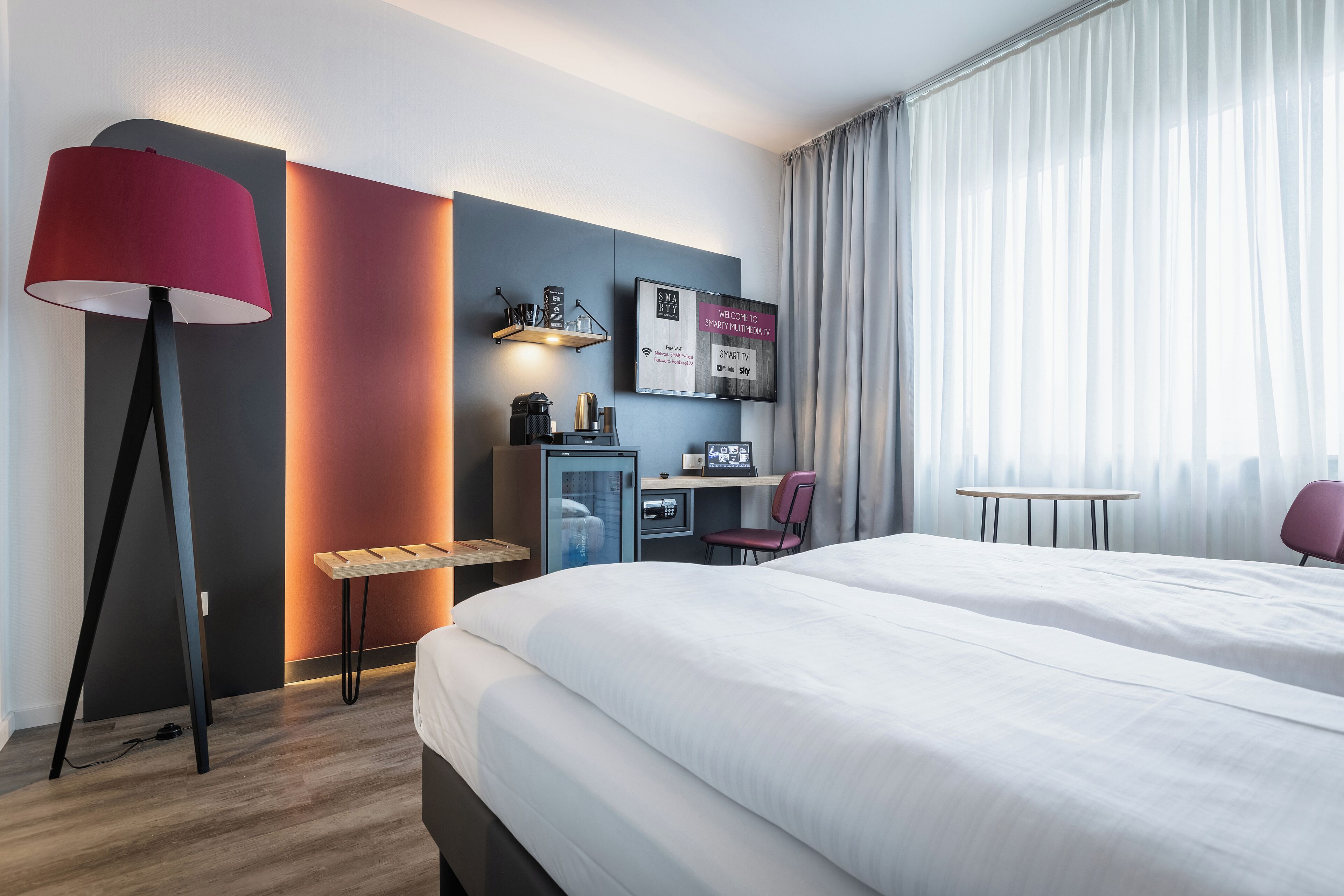 A hotel room with a bed, desk, chair, lamp, TV, and curtains.
