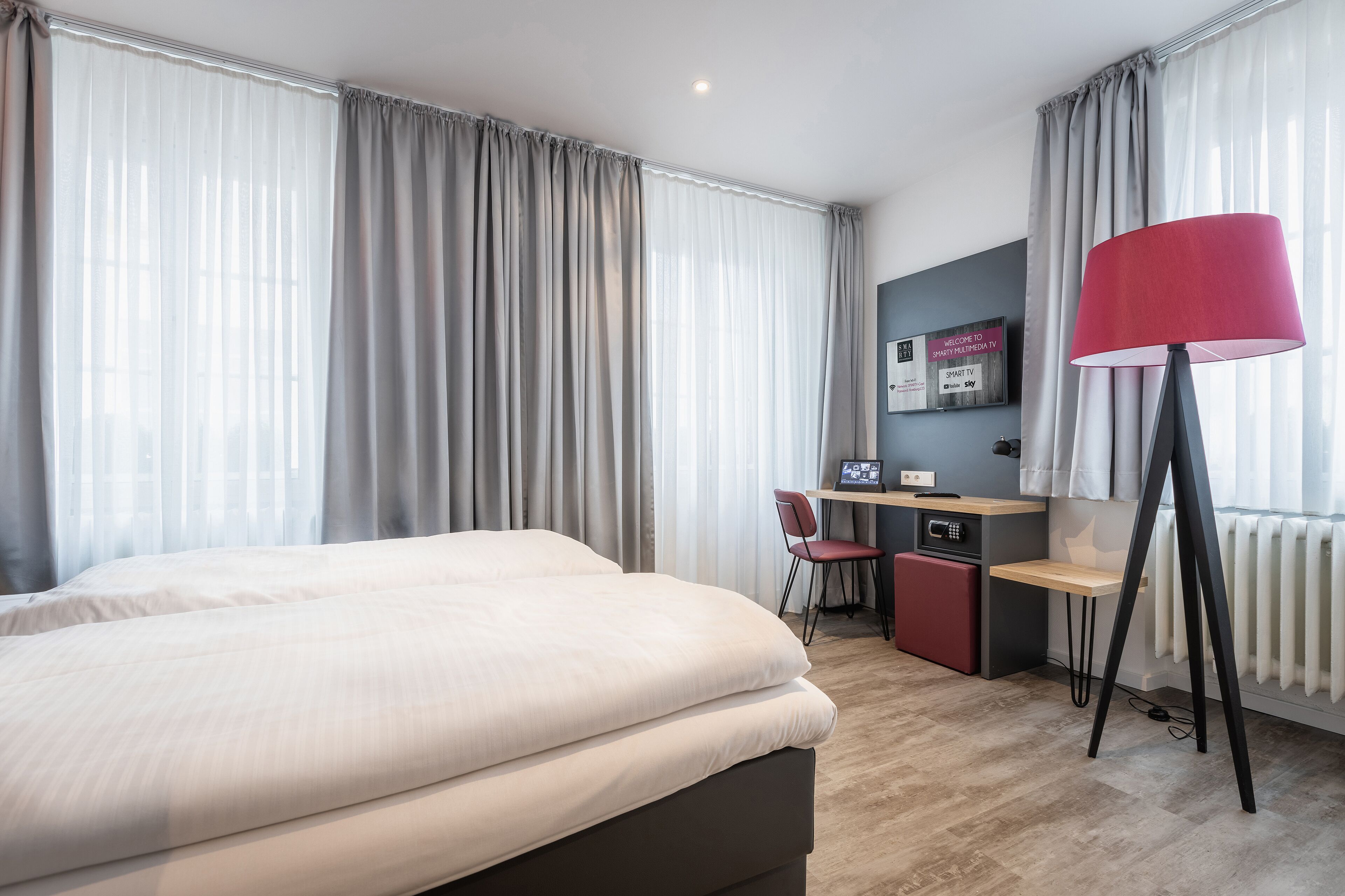 Habitación de hotel con una cama grande, un escritorio con computadora, una silla y una lámpara roja en el suelo.