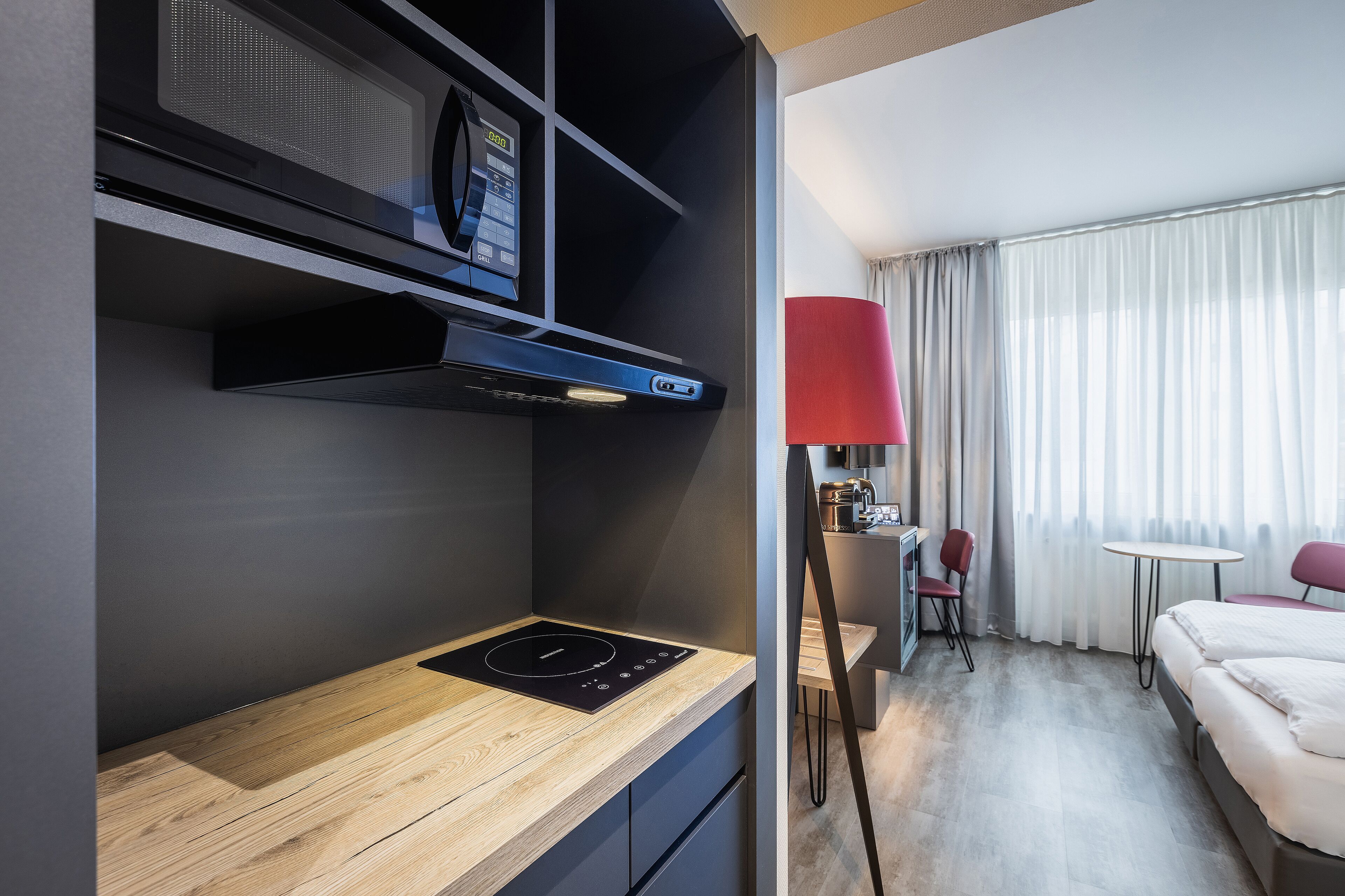 A modern hotel room with a built-in microwave, a compact kitchenette, a bed, a desk, and a red lamp.