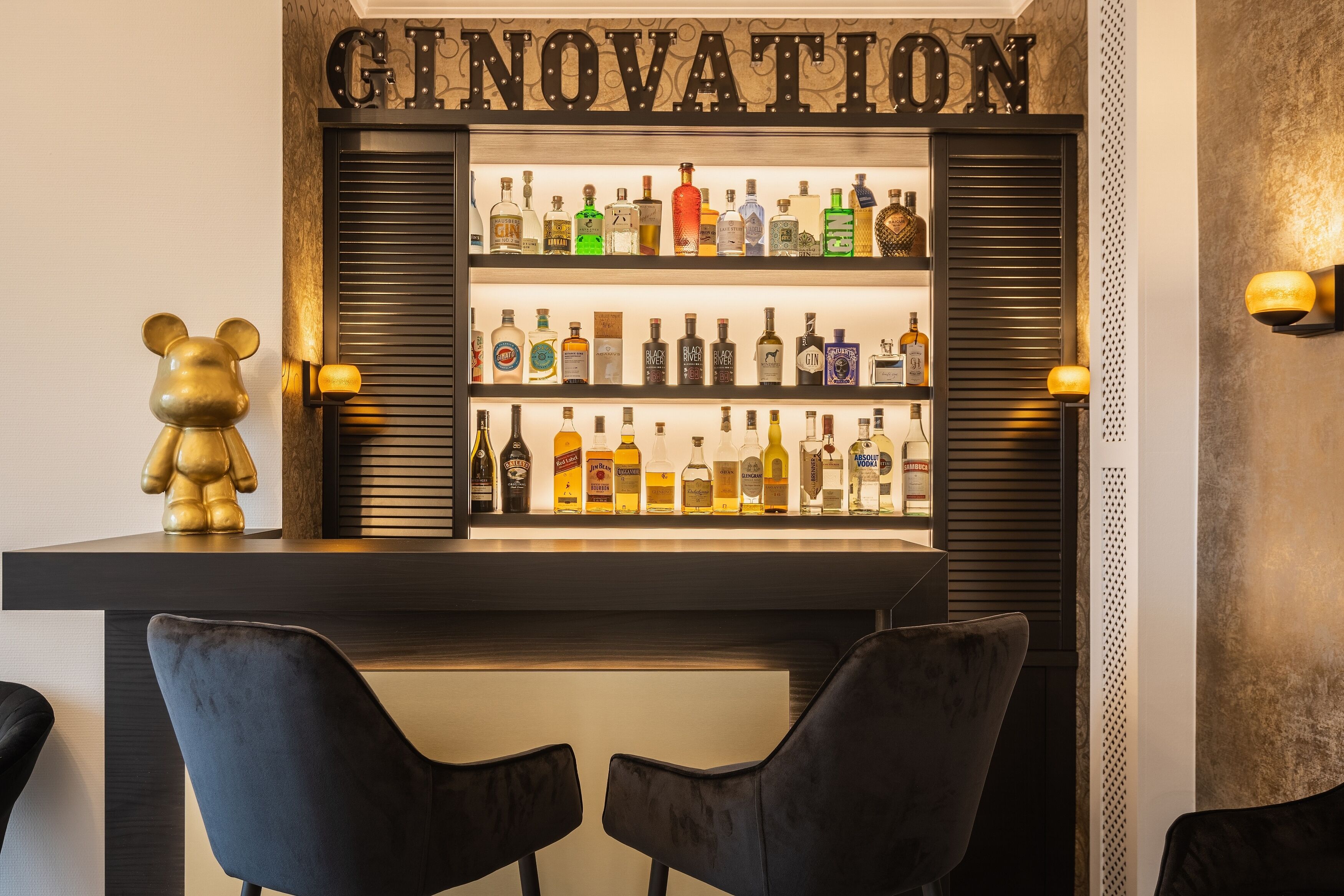 A bar area with shelves of liquor bottles, a bear figurine, and two black chairs.