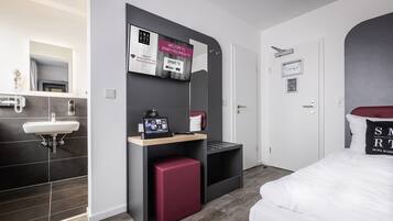 Una habitación de hotel moderna con una decoración en gris y blanco, un espejo grande, un televisor montado, un otoman rojo y una cama blanca.