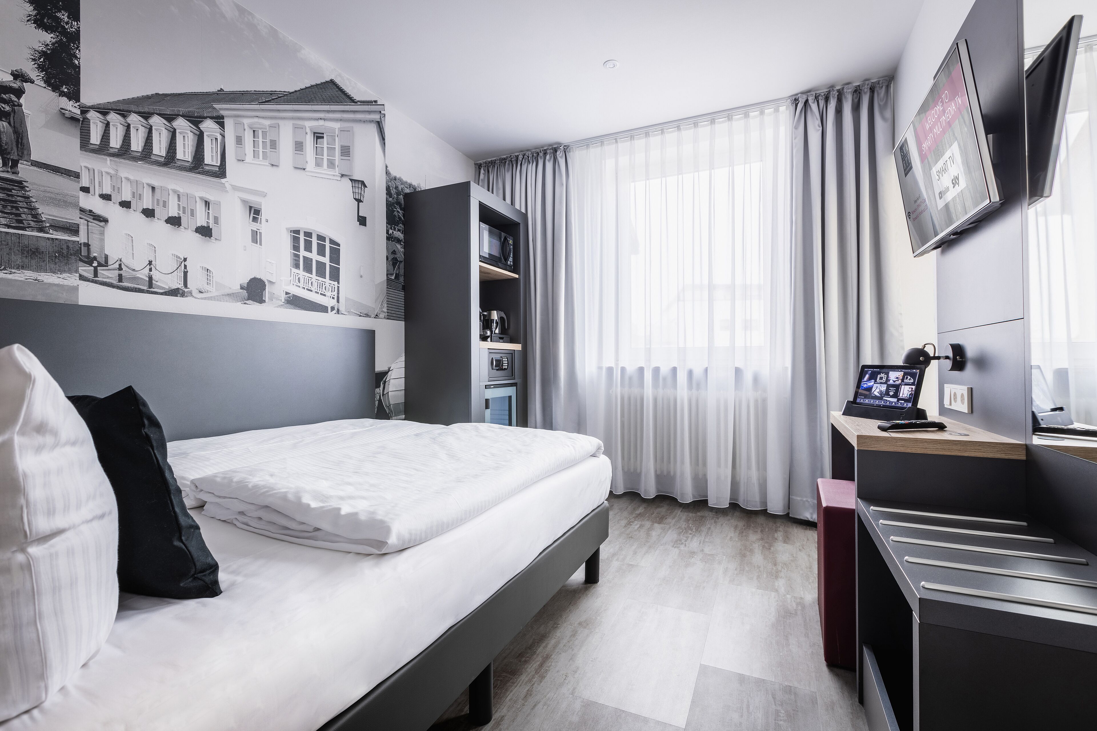 A modern hotel room with a neatly made bed, a flat-screen TV mounted on the wall, and a large window with sheer curtains.
