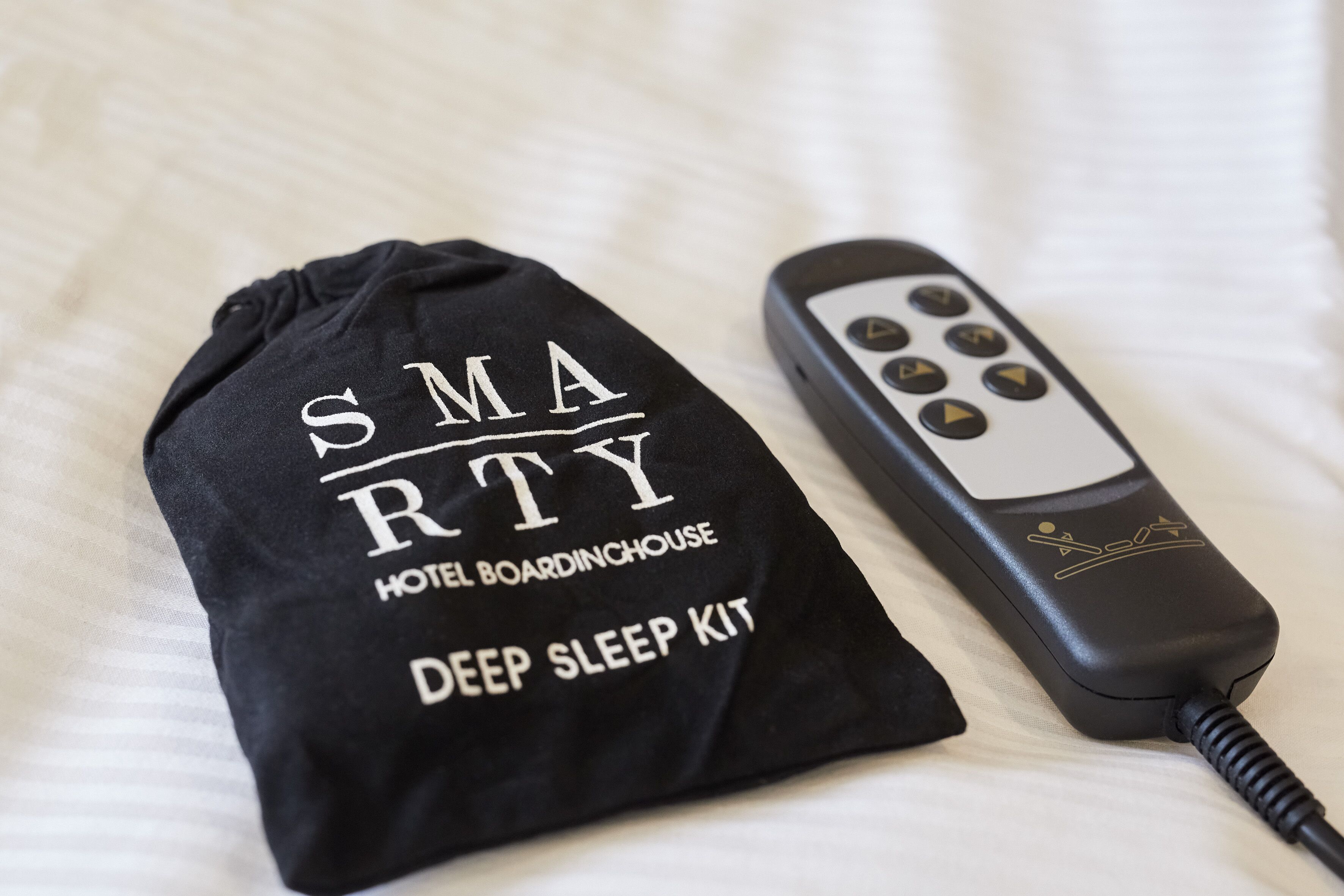 Un almohadón negro con la inscripción "SMARTY HOTEL BOARDINGHOUSE DEEP SLEEP KIT" y un control remoto negro.