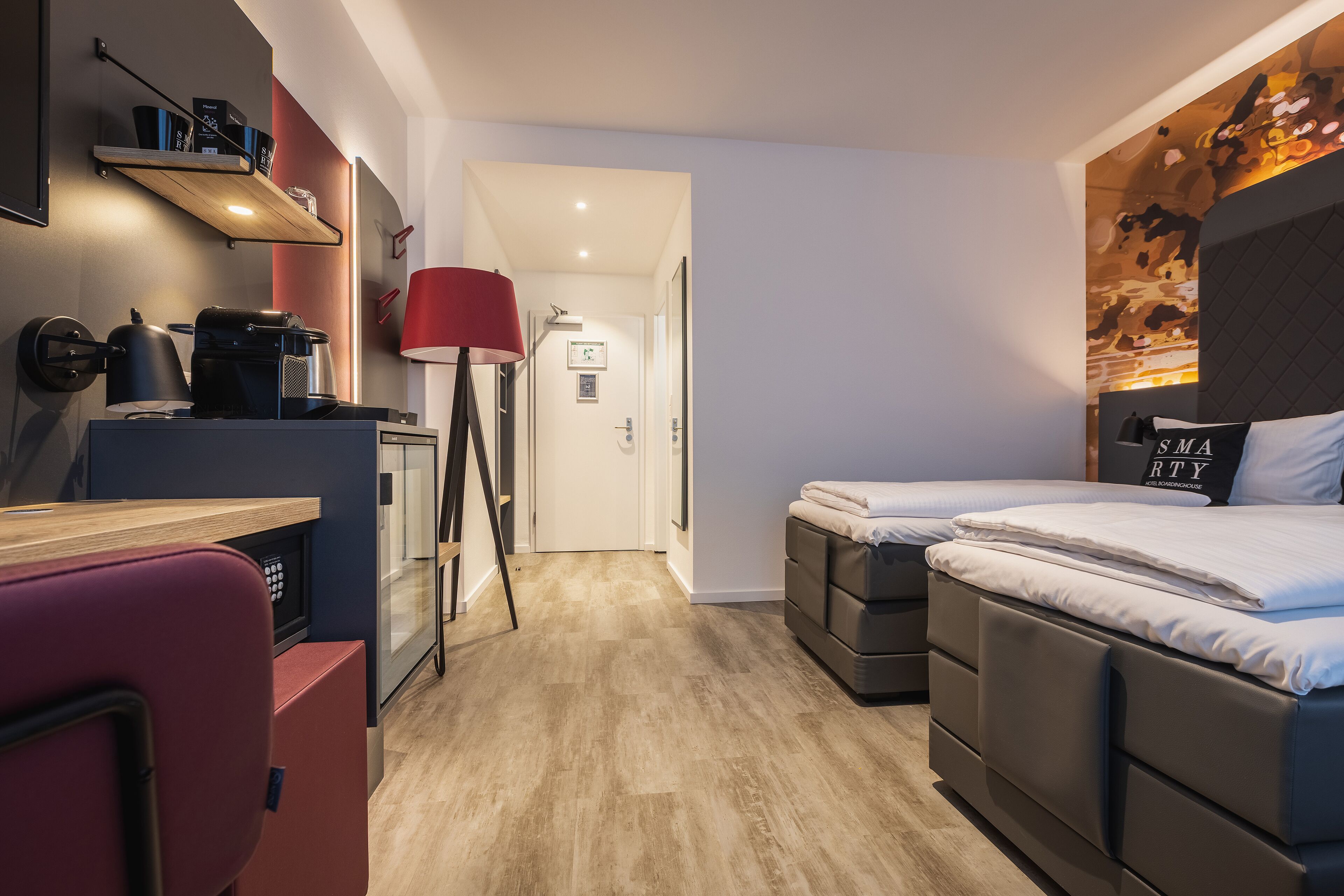 Une chambre d’hôtel compacte avec une kitchenette, un lit agrémenté d’un oreiller sur lequel est brodé le mot « SMARTY », et une lampe de sol rouge.