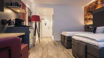 Une chambre d’hôtel compacte avec une kitchenette, un lit agrémenté d’un oreiller sur lequel est brodé le mot « SMARTY », et une lampe de sol rouge.