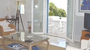 A living room with a glass coffee table, white sofa, and a dining area with white chairs. - Villa Amfitriti - Mykonos Guests 6 (Mykonos)