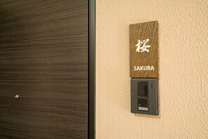 Un cartello di legno con caratteri giapponesi e la parola "SAKURA" sotto, fissato a una parete strutturata vicino a una porta.