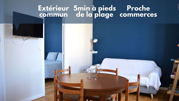 A room with a round wooden table and four chairs, a bed with a blue wall, a white sofa, and a TV mounted on the wall. - 2 steps from the beach - Centre d'Asnelles (Asnelles)