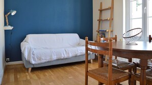 A room with a blue wall, a wooden floor, a white sofa, a wooden dining table, and a chair. - 2 steps from the beach - Centre d'Asnelles (Asnelles)
