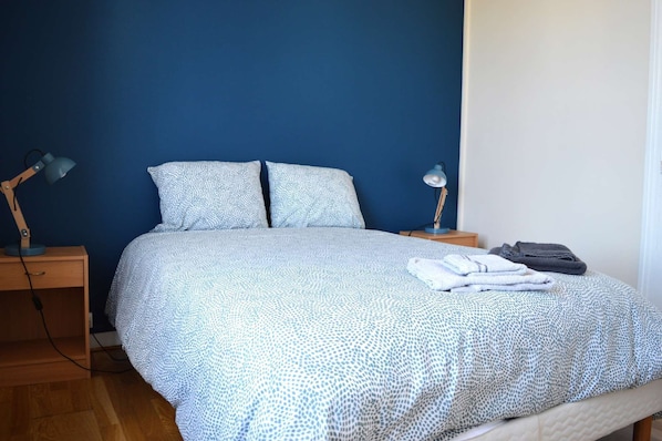 Una cama bien hecha con una colcha estampada, dos almohadas, una lámpara de noche y una toalla doblada sobre la mesita.