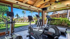 Un gimnasio con máquinas de cardio, un ventilador de techo y vistas a un resort con palmeras.