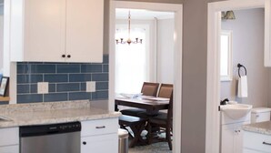 A kitchen with white cabinets, a blue backsplash, a dishwasher, and a dining area with a table and chairs. - Spacious Cityside House w/ Game Room & Fire Pit (Geneva)