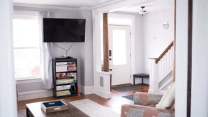 A living room with a flat-screen TV mounted on the wall, a bookshelf, a coffee table, and a patterned armchair. - Spacious Cityside House w/ Game Room & Fire Pit (Geneva)