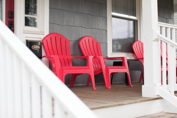 En veranda med tre røde Adirondack-stole, et lille bord, et hvidt gelænder og en grå facade.