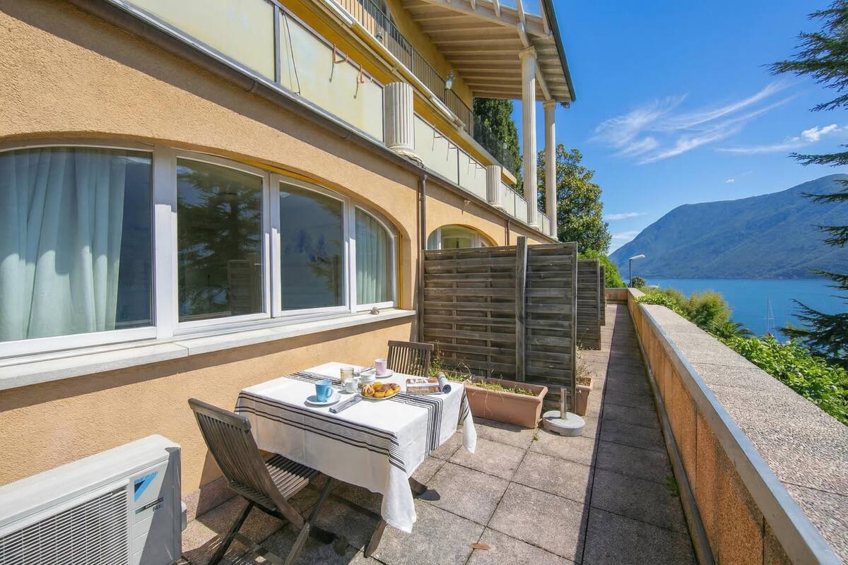 A terrace with a table set for two, a potted plant, and a view of a lake and mountains.