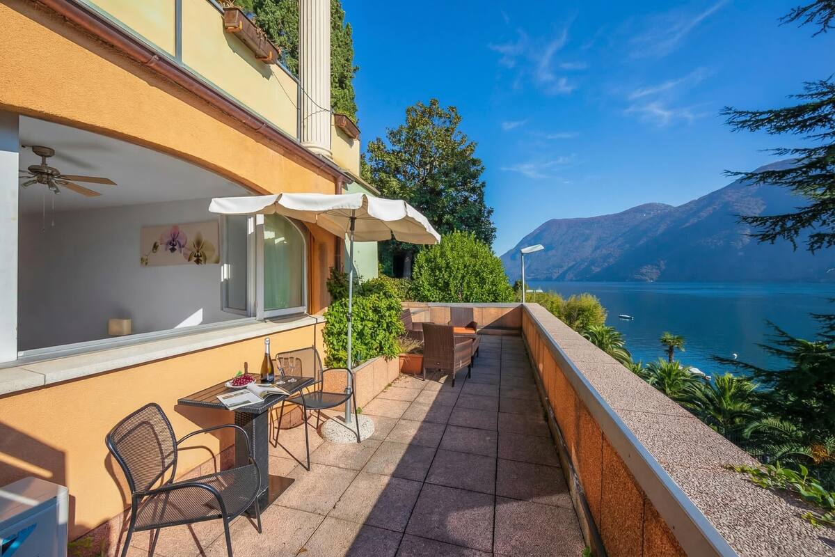A balcony with a table, chairs, and a white umbrella, overlooking a lake and mountains.