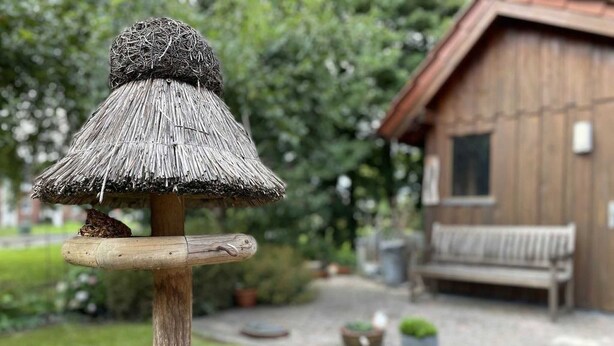 Eine rustikale Holzbank, ein reetgedecktes Vogelhaus und ein Gebäude mit Schindeldach inmitten eines Gartens.