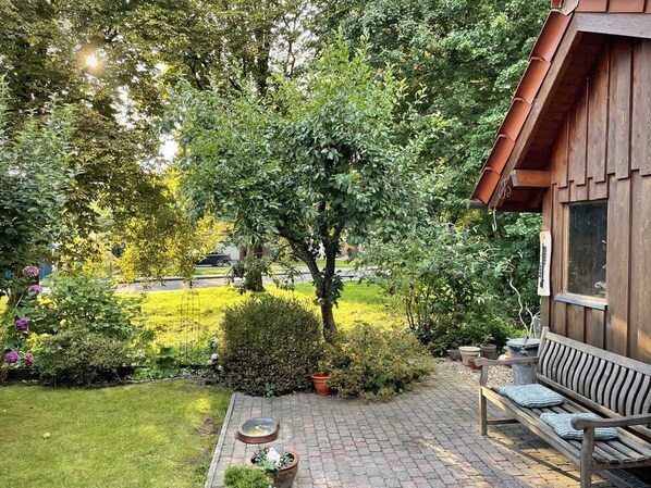 A wooden cabin with a brick patio, surrounded by lush greenery and a garden with flowers and shrubs.