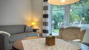 Ein Wohnzimmer mit einer Couch, einem runden Tisch mit gemustertem Tischläufer, einer Lampe und Blick ins Grüne.