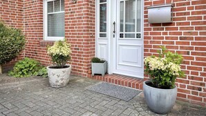 Ein Ziegelhaus mit zwei weißen Türen, einer kleinen Veranda und zwei großen Kübelpflanzen.