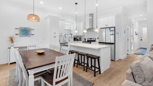 A modern kitchen with white cabinets, a wooden dining table, and pendant lighting. - ⭐6 Bedroom Beauty-Fab Interior-Great Outdoor Living Space-6 Bikes-6BR Cozy Shore (Miramar Beach)