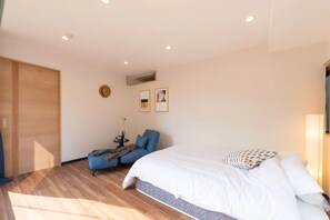 A modern hotel room with a large bed, a blue armchair, a small table with a vase, and a wooden wardrobe.