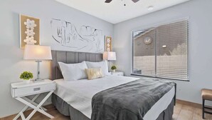 Een moderne slaapkamer met een groot bed, twee nachtkastjes met lampen, een kunstwerk aan de muur en een raam met zonwering.