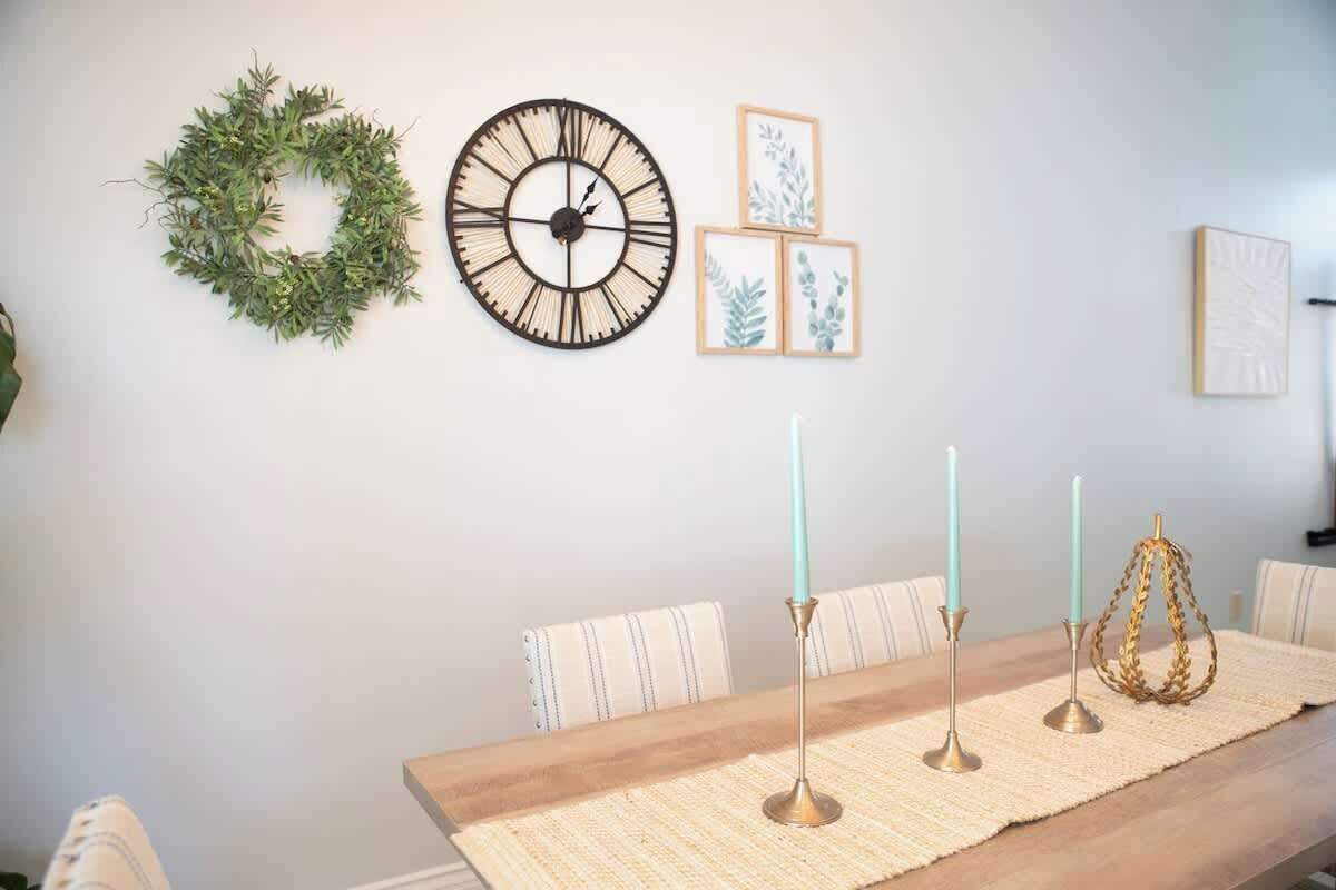 Een eetruimte met een houten tafel, gestreepte stoelen, een geweven placemat, een wandklok, ingelijste botanische prints en een krans.