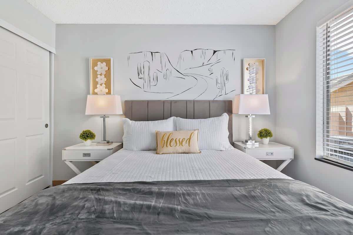 Un dormitorio con una cama, dos mesitas de noche con lámparas, una obra de arte en la pared y una ventana con persianas.