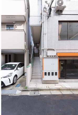 A narrow alleyway with a white car parked on the left, a building with a tiled facade, and a utility box on the right.